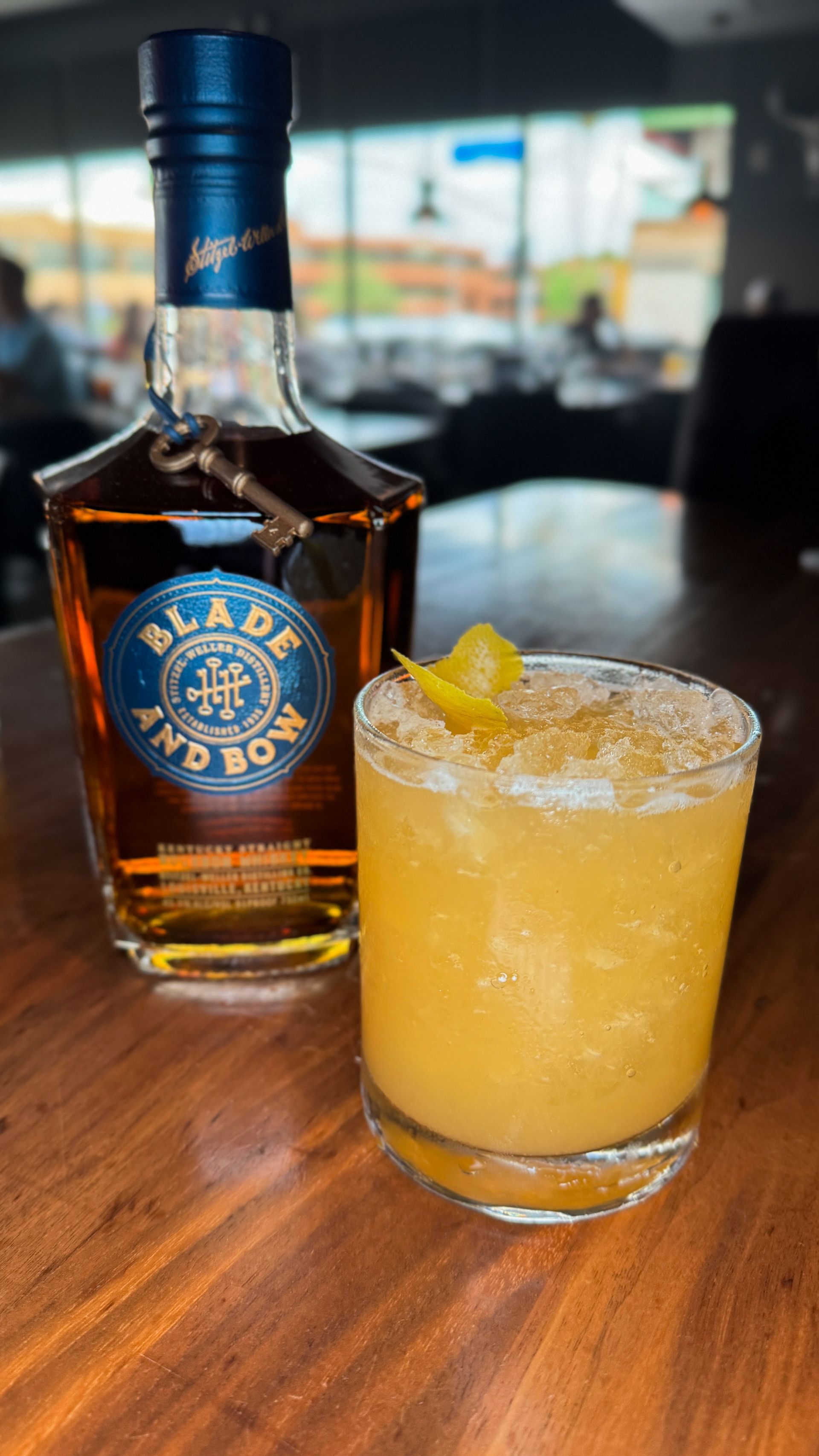 Blade and Bow whiskey bottle and cocktail on a wooden table. Yellow drink with lemon peel garnish.