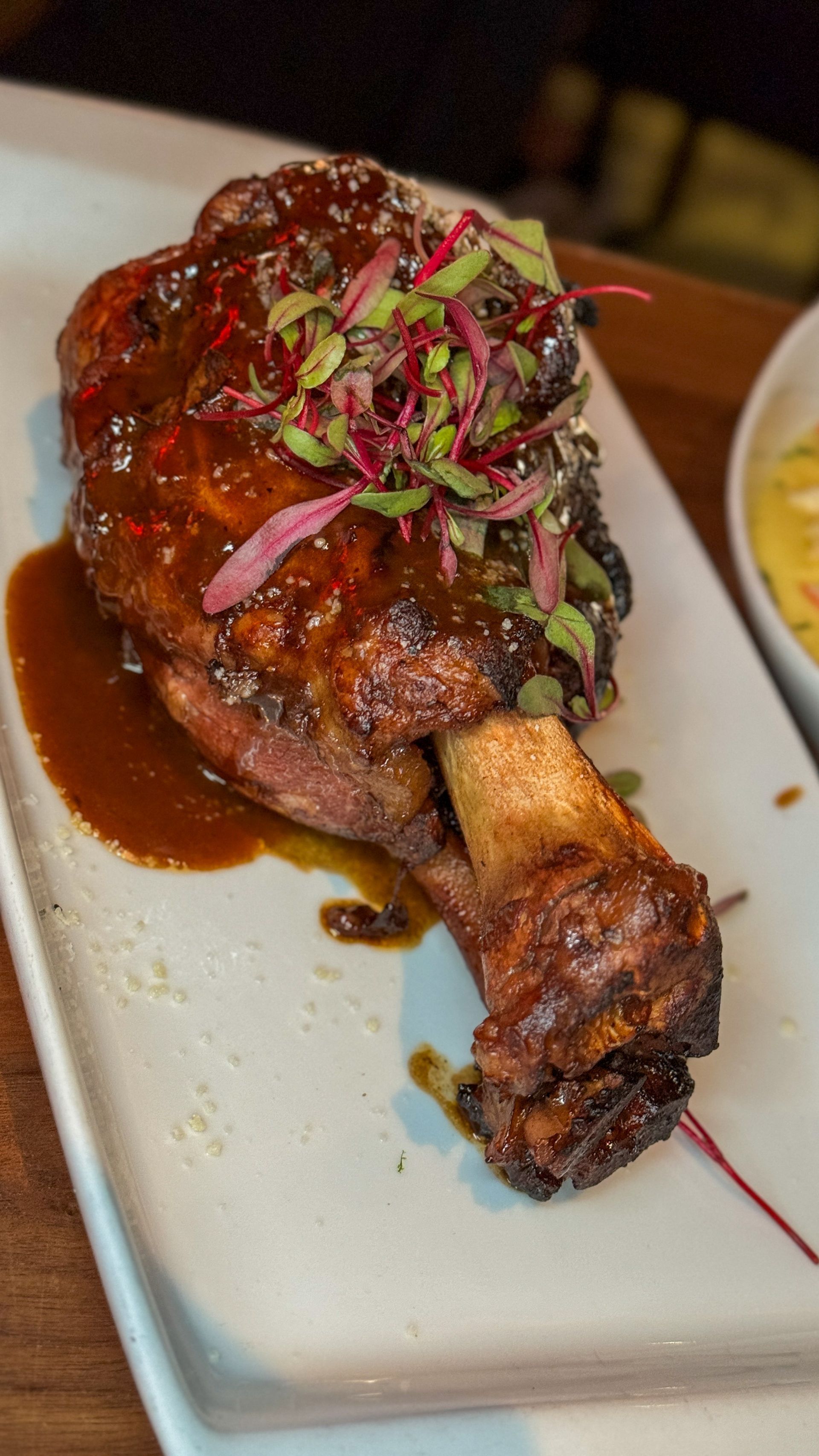 Braised meat on bone, drizzled with sauce and topped with red garnish, on a white rectangular plate.