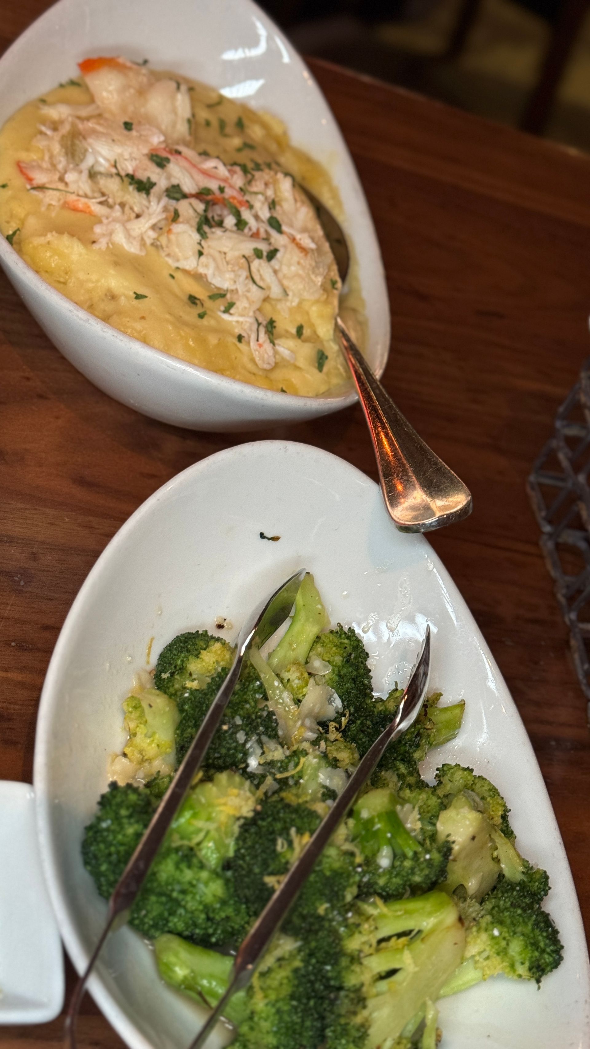 Two white oval dishes of food on a wooden table. One has crab dip, the other has broccoli.