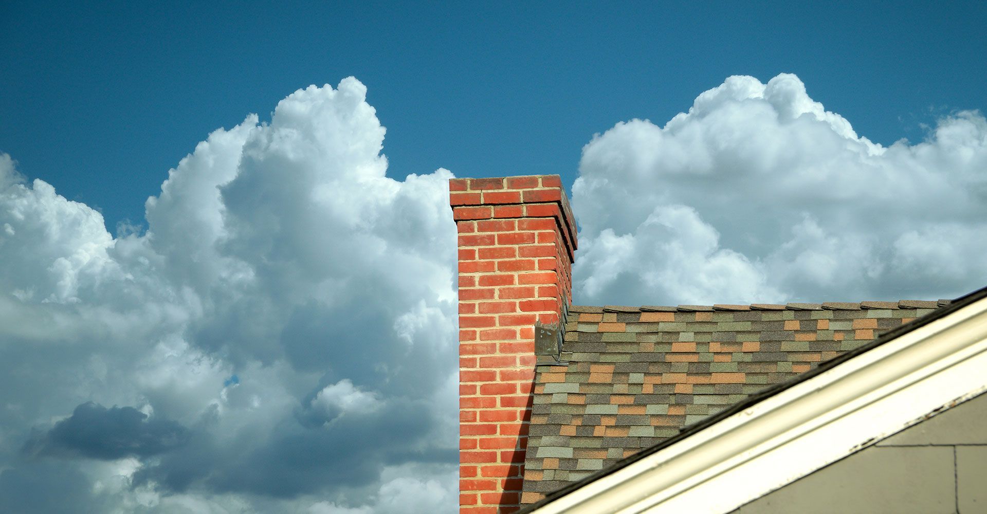 Brick chimney atop a house roof against a backdrop of puffy white clouds in a blue sky.