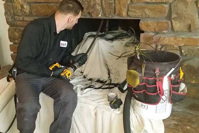 Chimney sweep cleaning a fireplace, using tools. Black clothing, white sheet, stone fireplace.