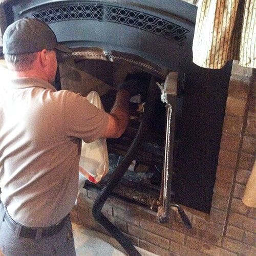 Man cleaning a fireplace with a vacuum; interior shot with brick and dark metal.