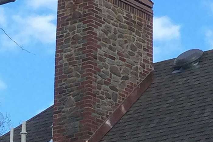 Brick and stone chimney on a dark shingle roof with a blue sky background.