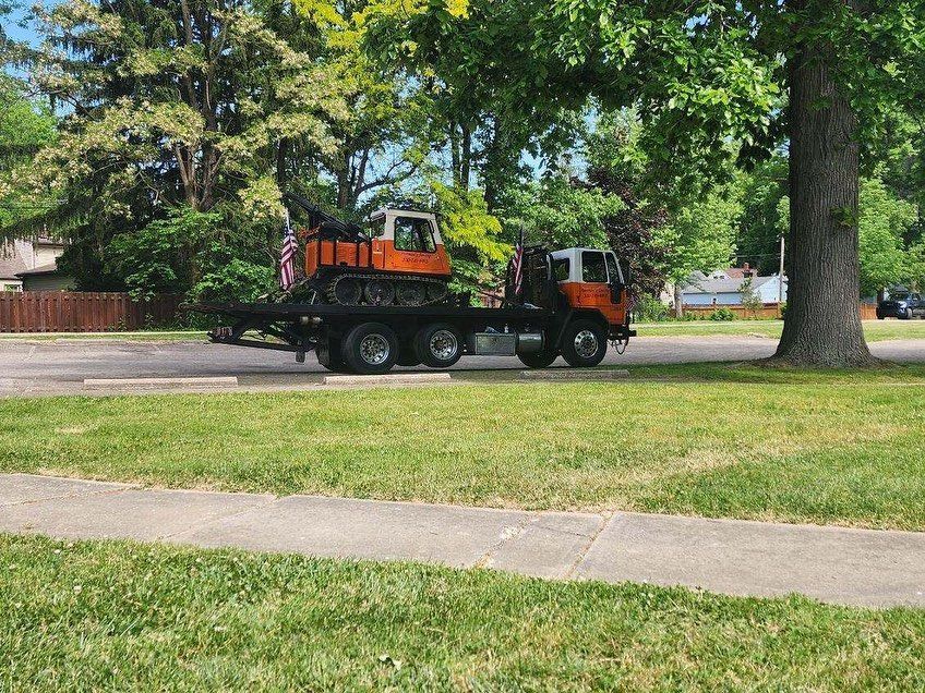 Truck with orange excavator on a trailer parked next to a tree in a residential area.