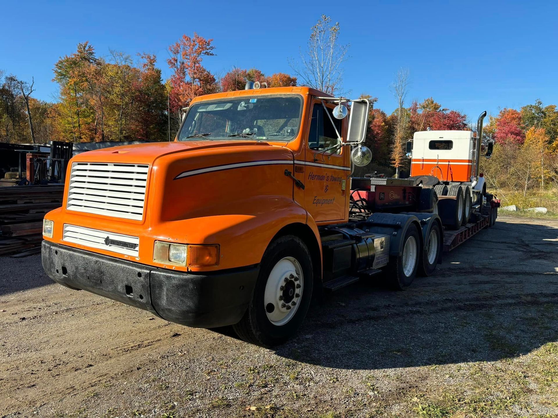 Orange semi-truck on a gravel lot; another truck is on its trailer. Fall foliage and blue sky in the background.