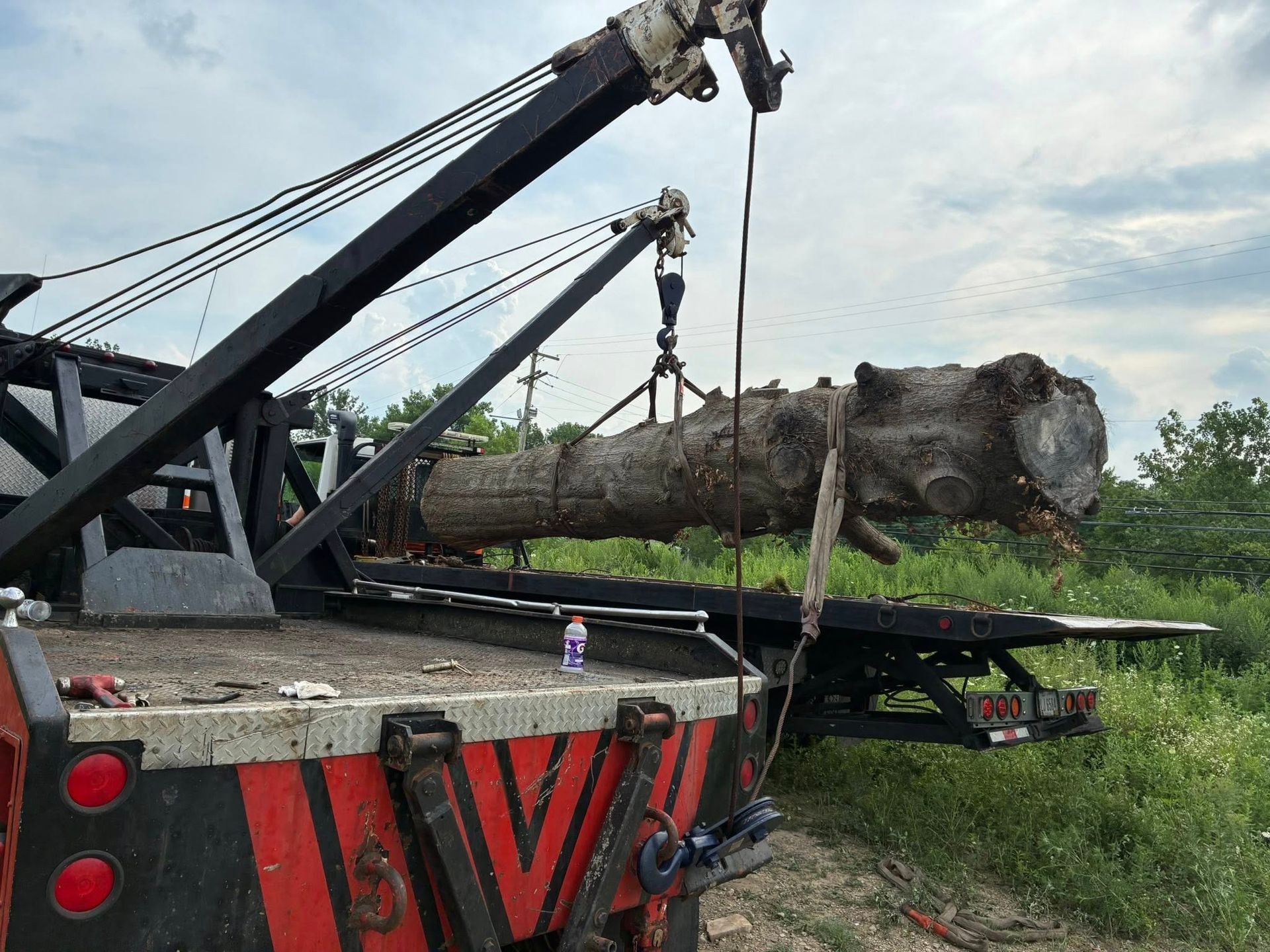 A tow truck lifting a large tree trunk. The truck is red and black, in a field, under a cloudy sky.