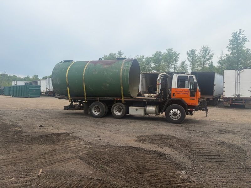 Orange flatbed truck carrying a large green tank in a dirt lot