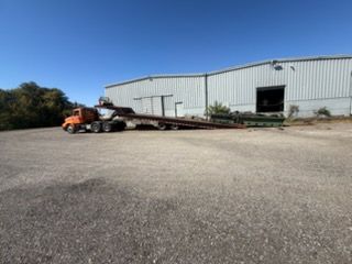 Orange forklift with a long trailer in front of a large metal warehouse on a gravel lot