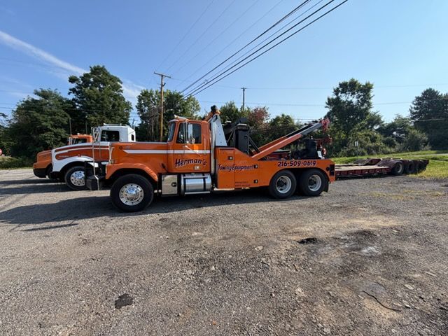 Orange tow truck parked on gravel roadside under power lines, with a long boom and flatbed.
