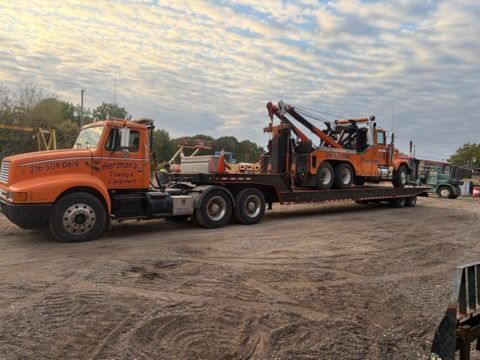 Orange tow truck hauling a heavy orange recovery vehicle on a flatbed in a dirt lot under cloudy sky