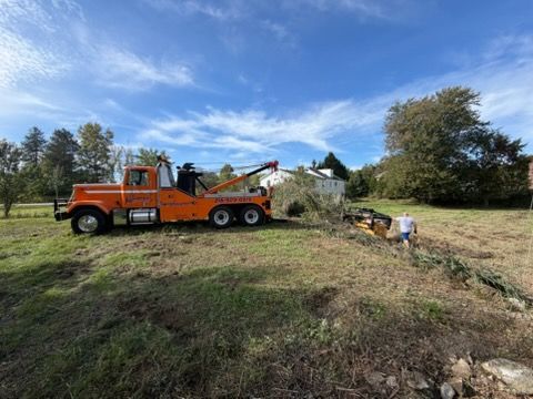 Orange tow truck working on a grassy roadside near two people and a fallen tree under a blue sky