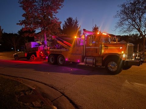 Tow truck with flashing lights parked on a street at dusk, illuminated by red and amber lights