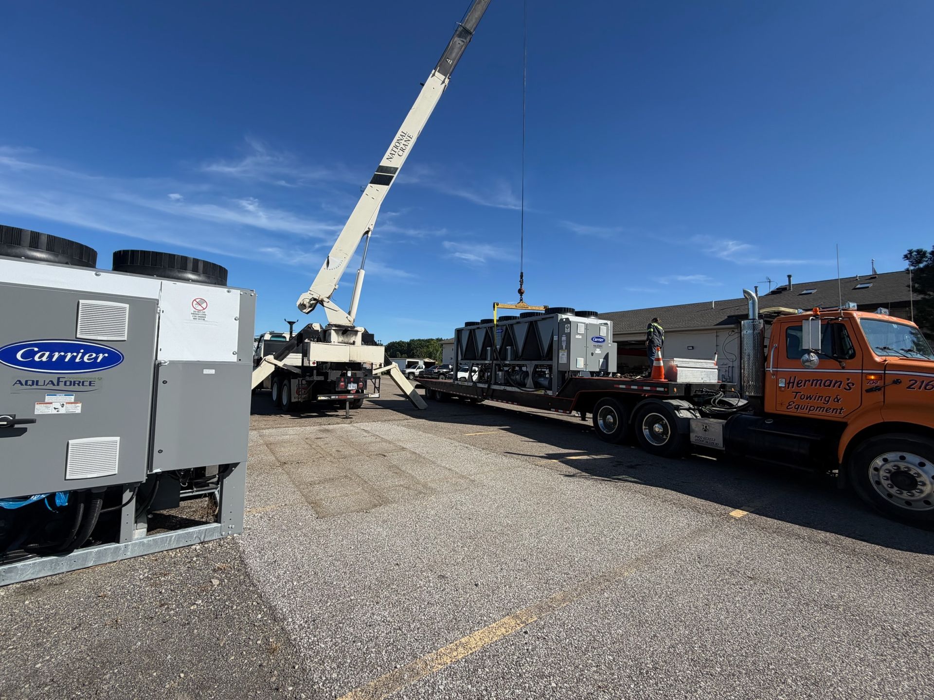 Crane lifting equipment onto flatbed trucks in a gravel lot under a blue sky
