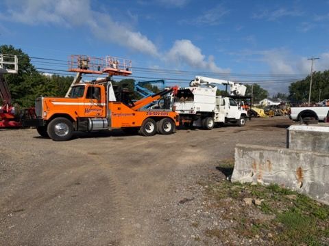 Tow truck loading a heavily damaged black car. A man secures straps. Sunny outdoor scene.