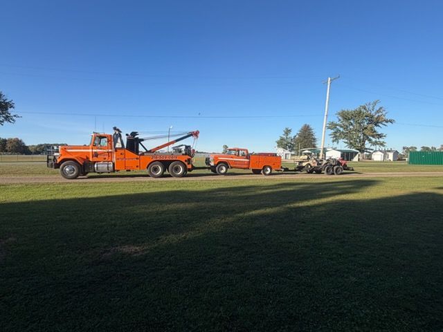 A tow truck lifts a large tree trunk, with a worker atop the truck, in a grassy field under a cloudy sky.