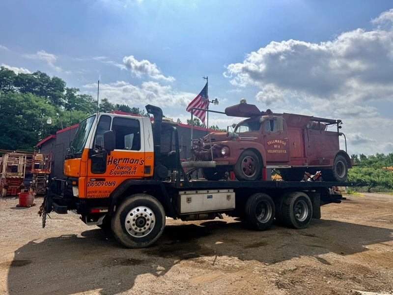 Orange tow truck carrying a red fire engine on a dirt lot under a cloudy sky