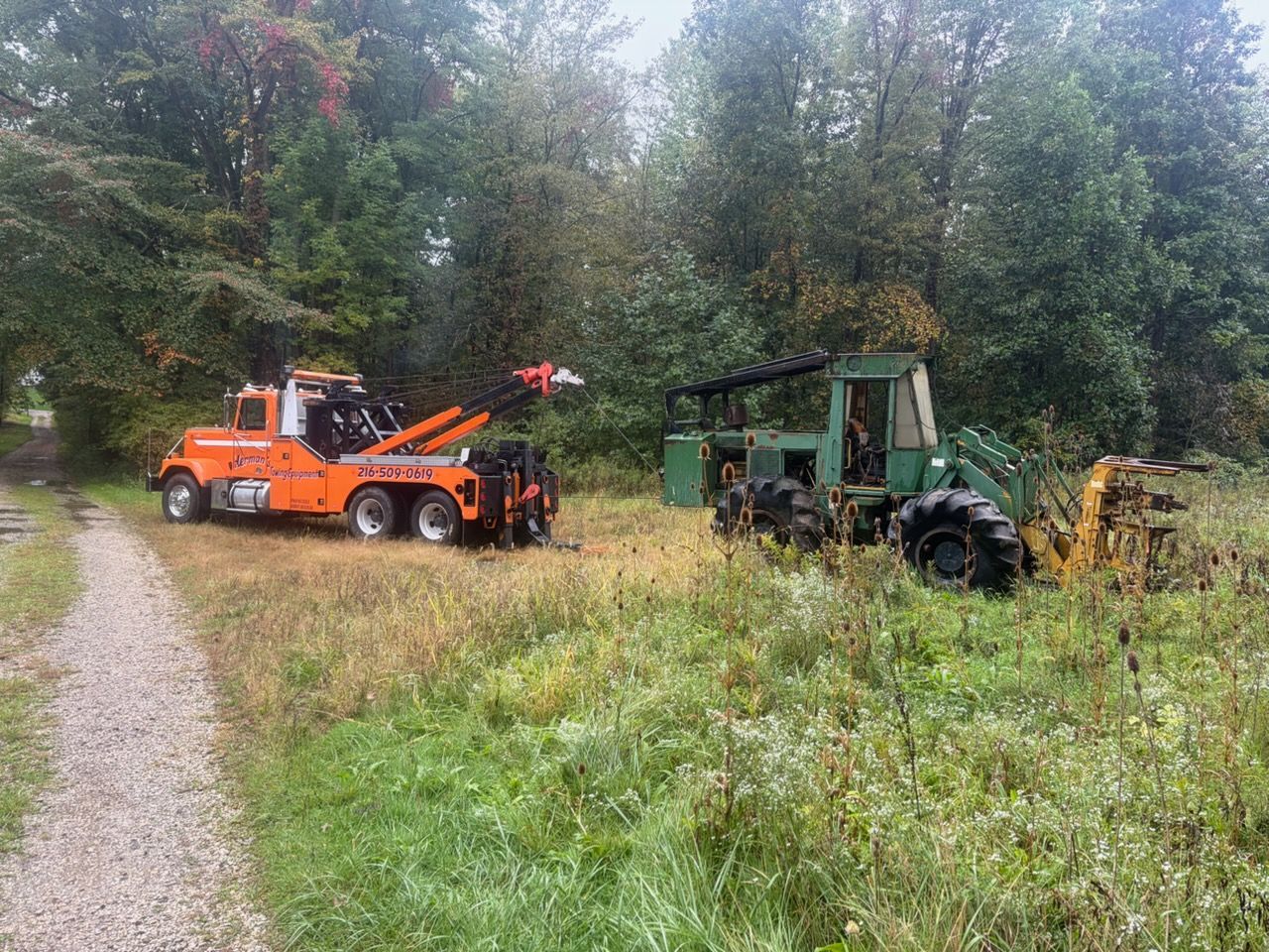 Orange tow truck and green tractor parked by a gravel path in a wooded field