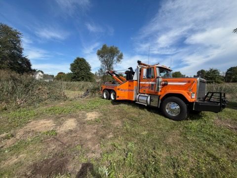 Orange tow truck with boom parked on a grassy dirt field under a partly cloudy sky