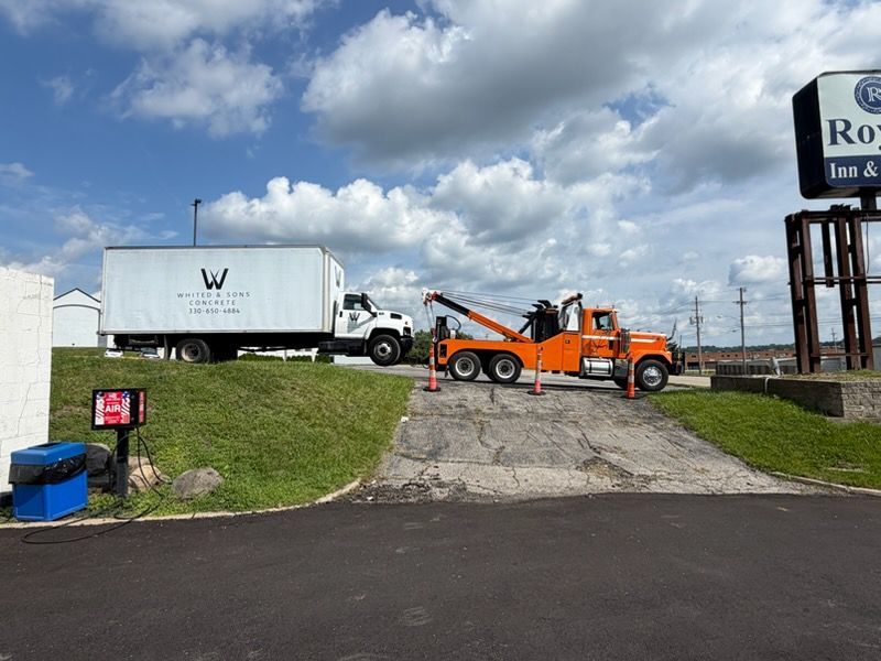 Tow truck hauling a white box truck on a driveway beside a roadside sign under cloudy skies