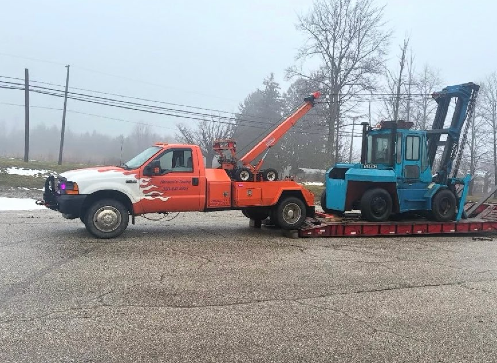 Orange tow truck hauling a blue forklift on a red flatbed trailer in a foggy roadside scene
