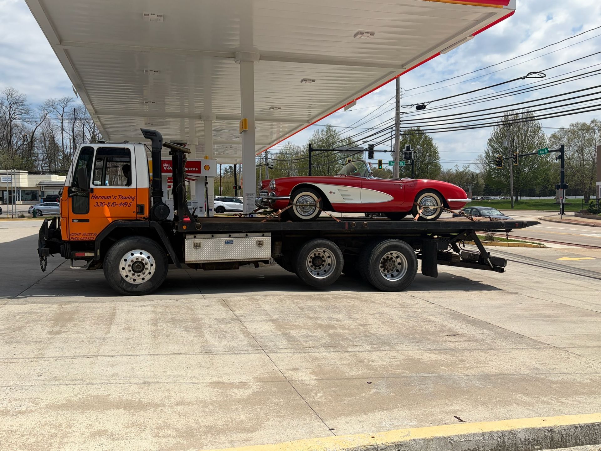 Orange tow truck carrying a red classic car at a gas station under a canopy