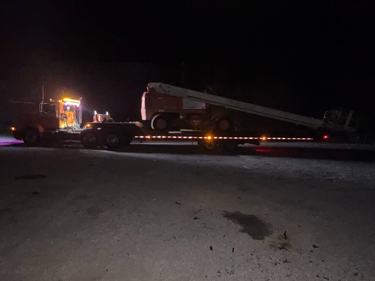 Tow truck towing a large utility vehicle at night on a dark road, with flashing lights.