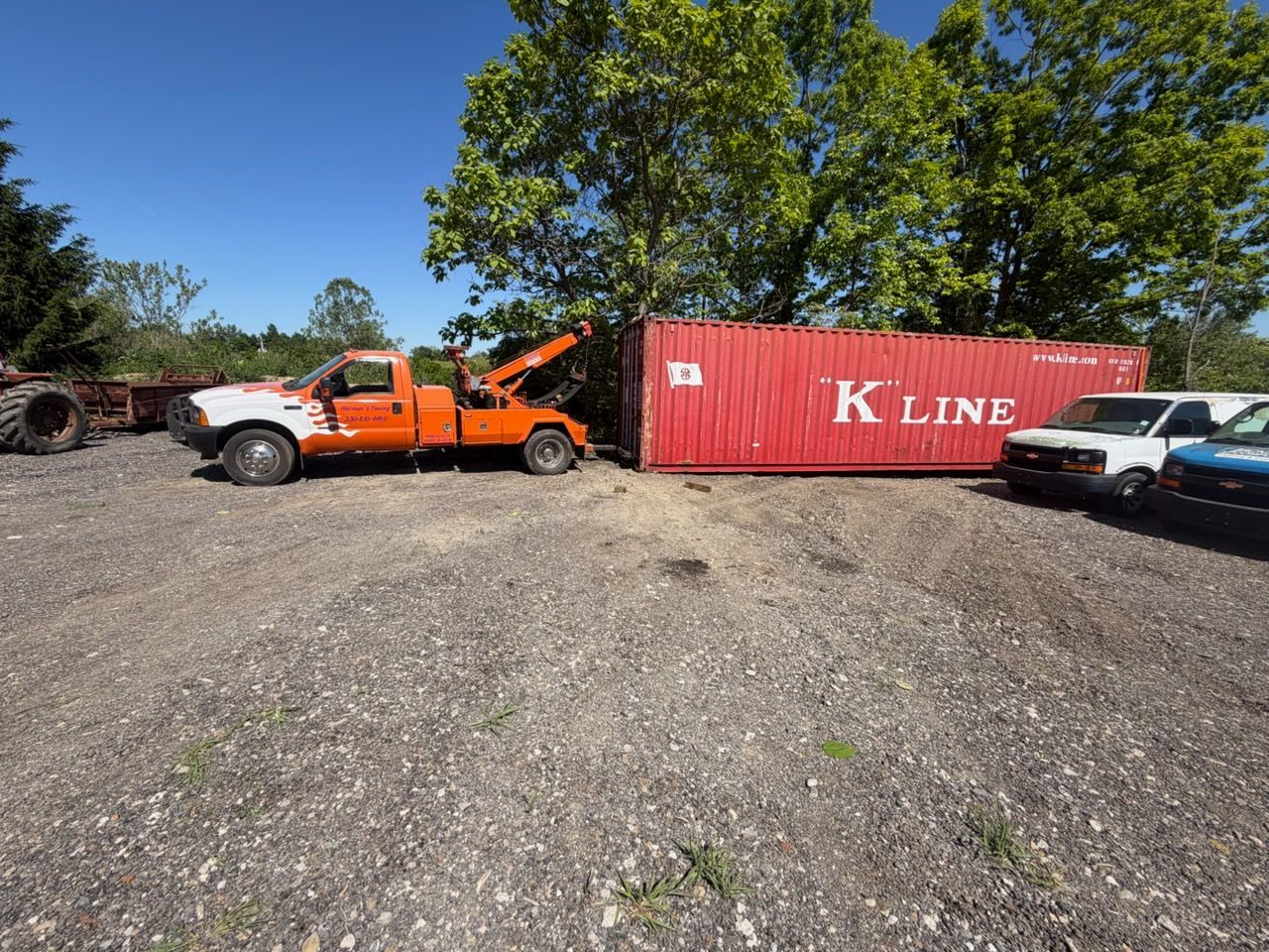 Orange tow truck beside red KLINE shipping container in a gravel lot under trees