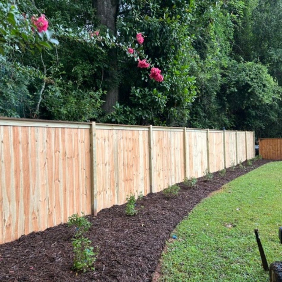 New wooden fence beside a mulched garden bed with small shrubs and a green lawn