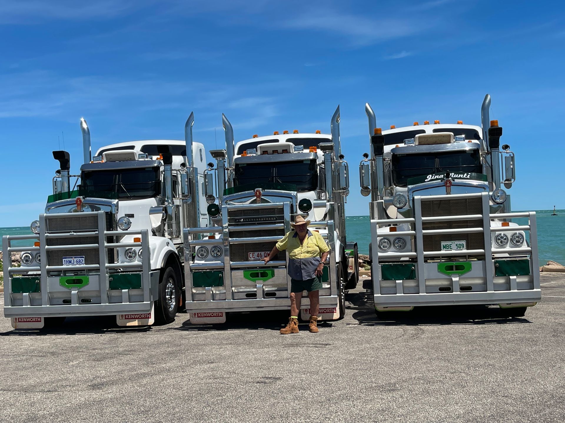 Man stands between three silver and green semi-trucks, under a bright blue sky by the water — Frontier Cement Supplies In East Arm, NT
