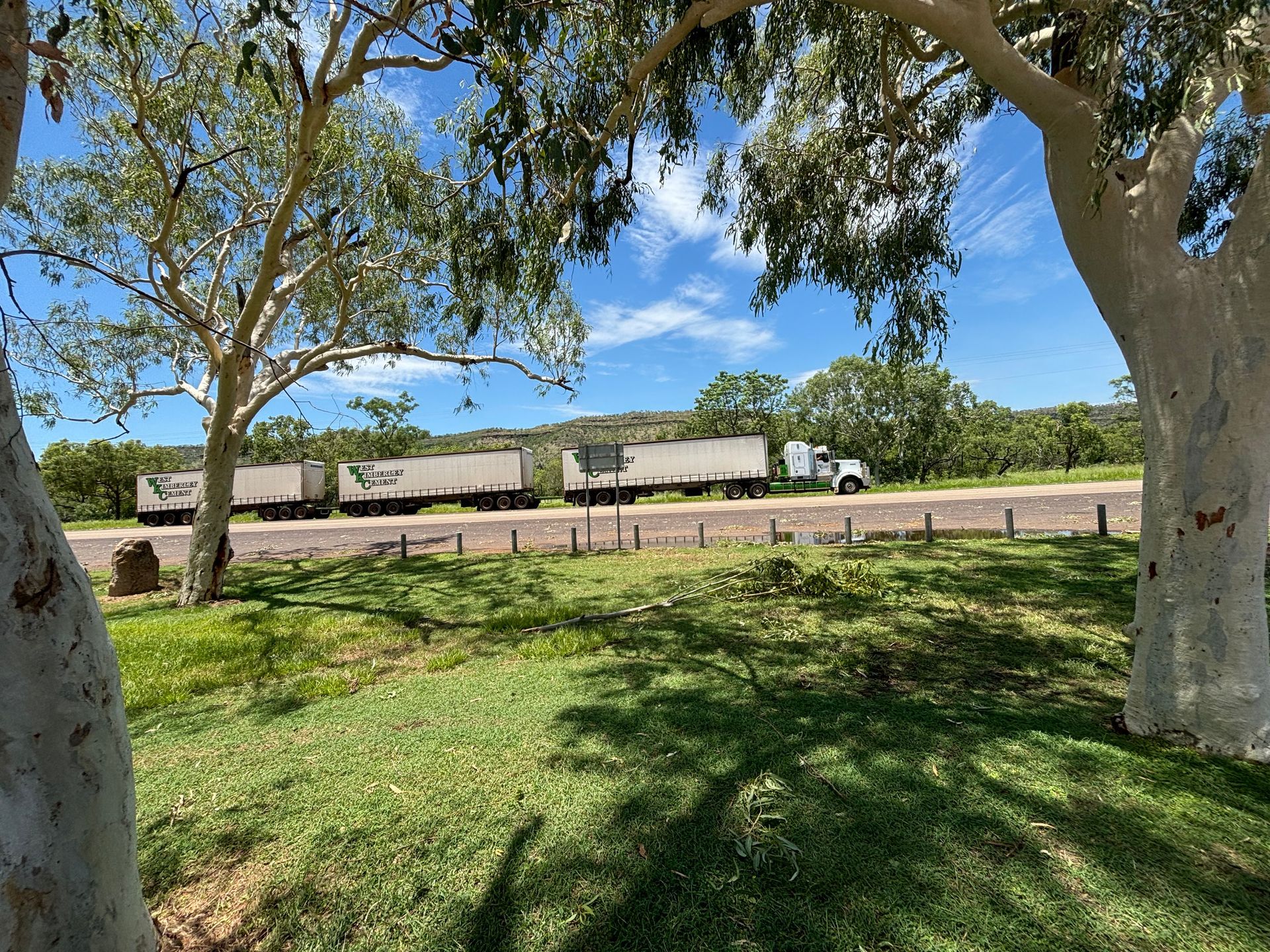 Shaded grassy park with large trees and a white train passing behind a fence under a blue sky — Frontier Cement Supplies In East Arm, NT