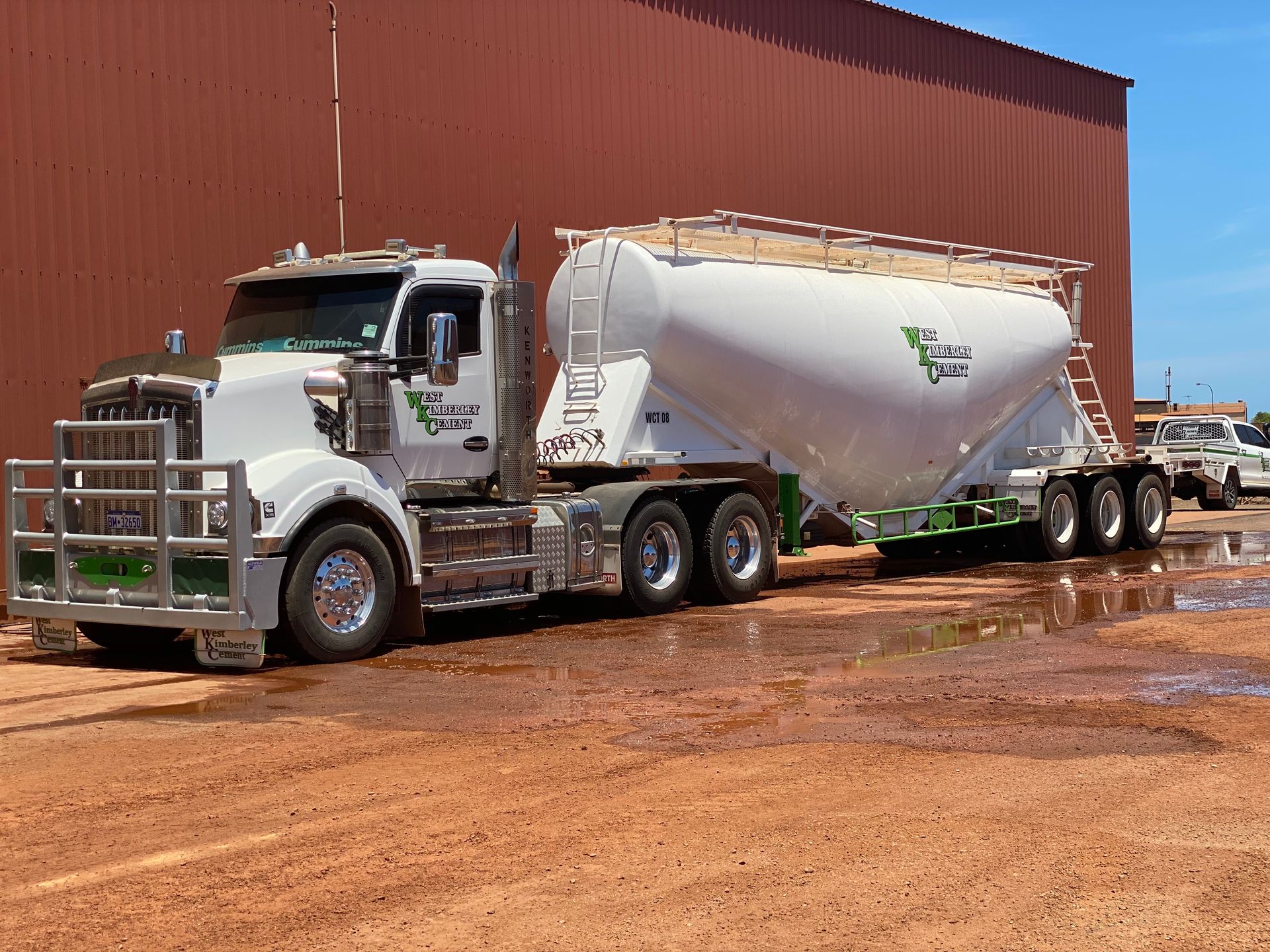 White semi-truck with a large white tanker trailer parked on reddish dirt near a brown building — Frontier Cement Supplies In East Arm, NT