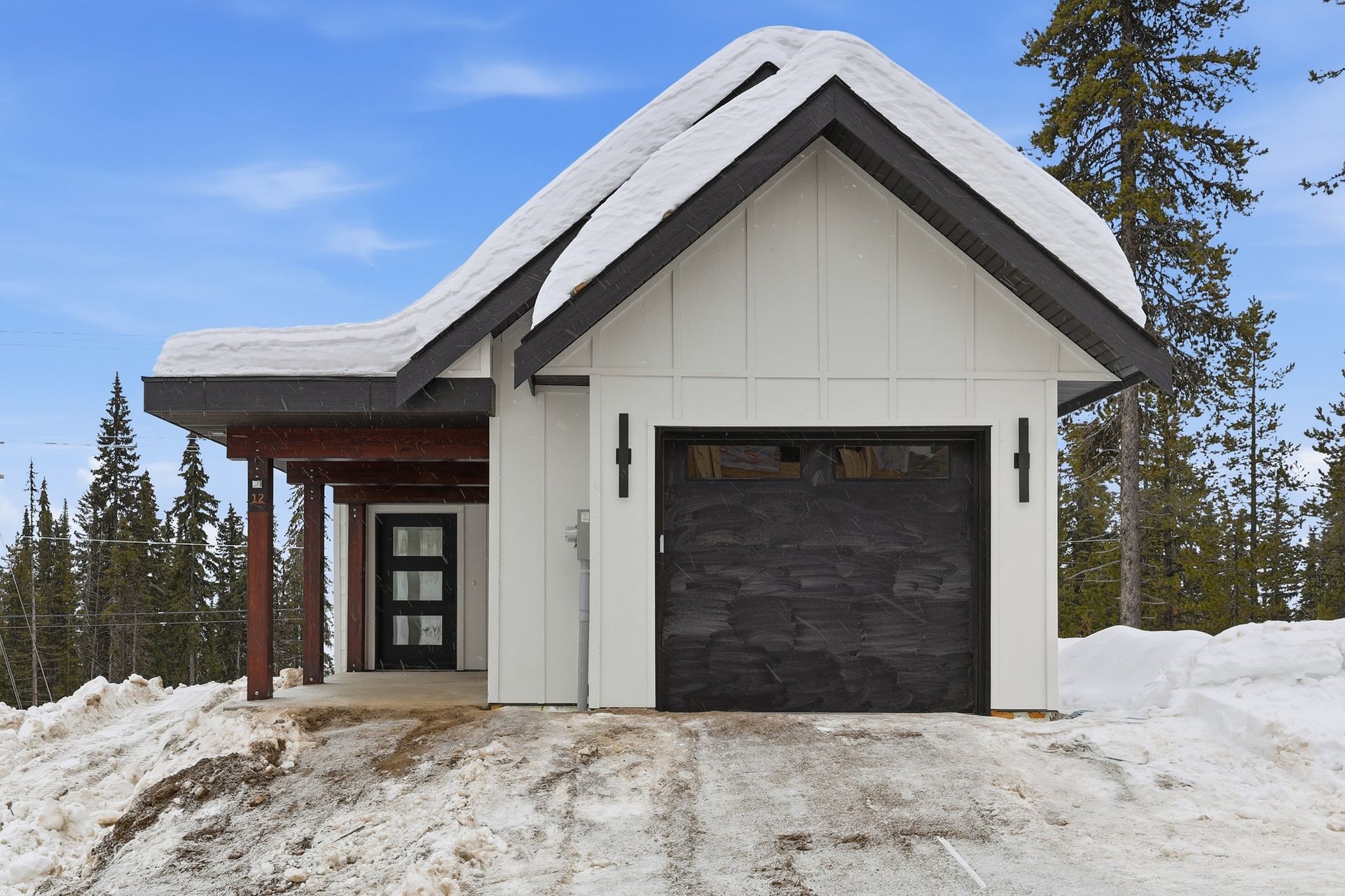 Snow-covered modern cabin with a dark garage door and tall evergreens in the background