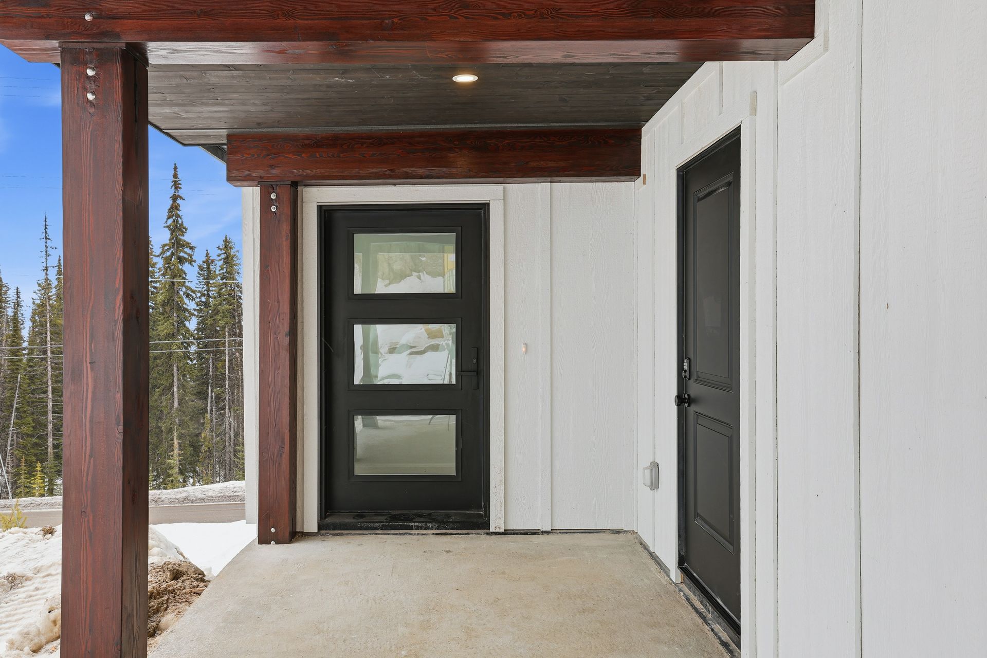 Covered porch with a black front door and white siding, overlooking snowy ground and pine trees.