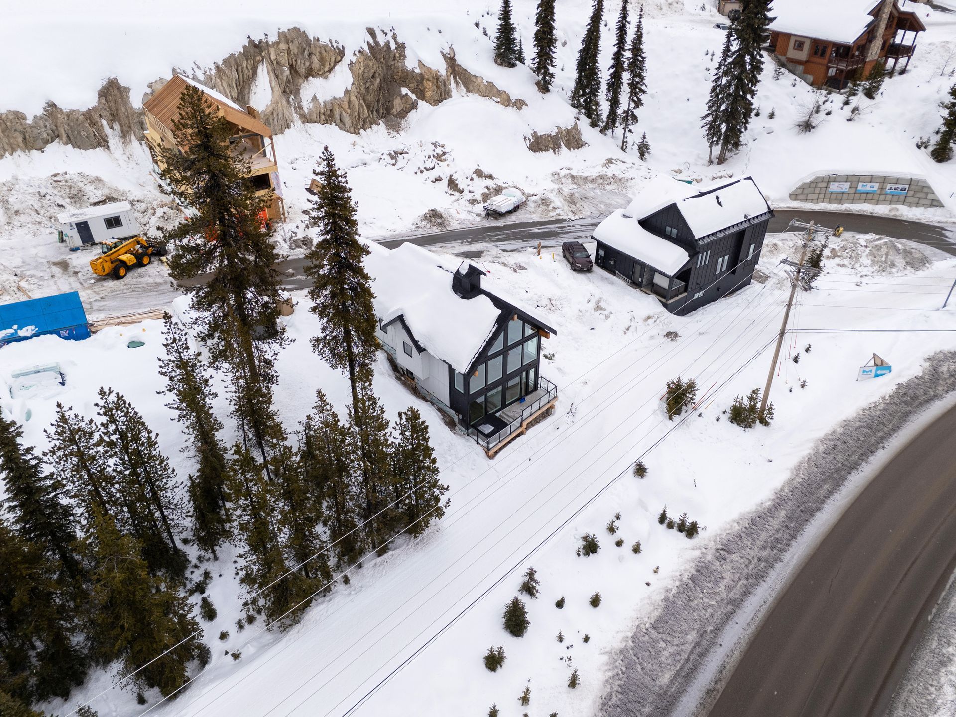Snow-covered mountain homes beside a winding road, with evergreen trees and parked vehicles.