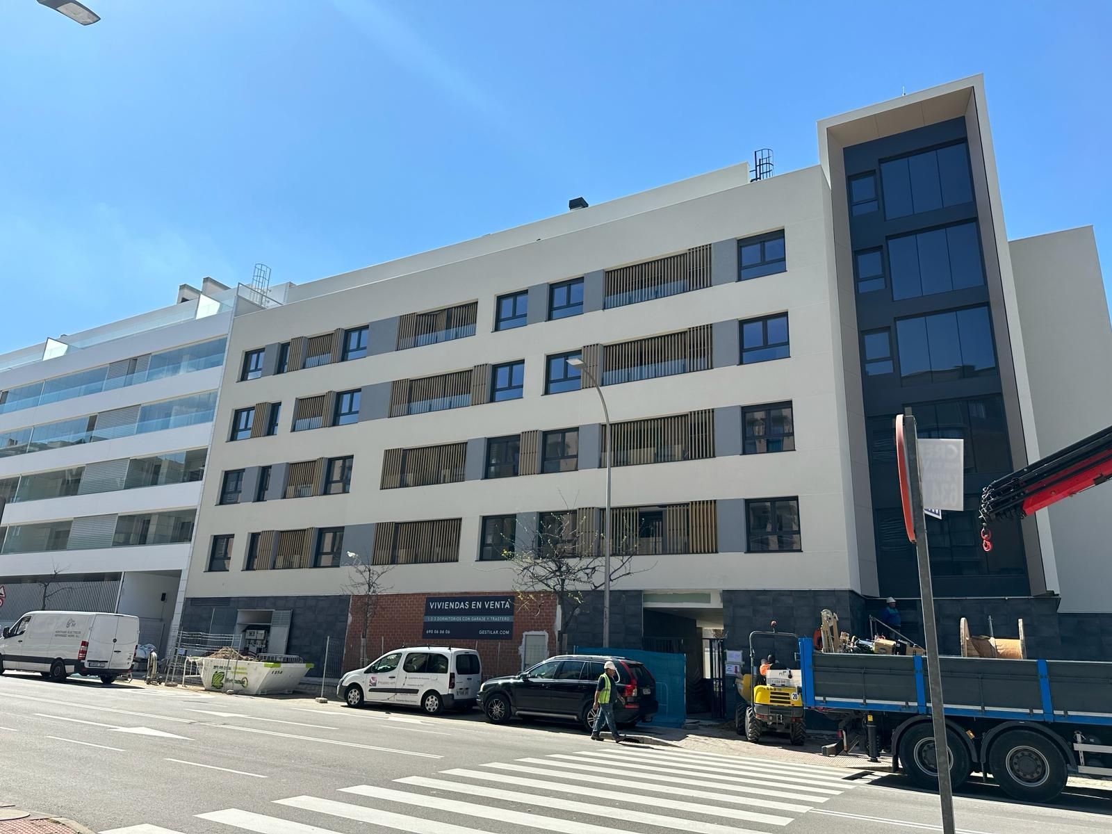 Moderno edificio de apartamentos en una calle soleada con coches aparcados y maquinaria de construcción.