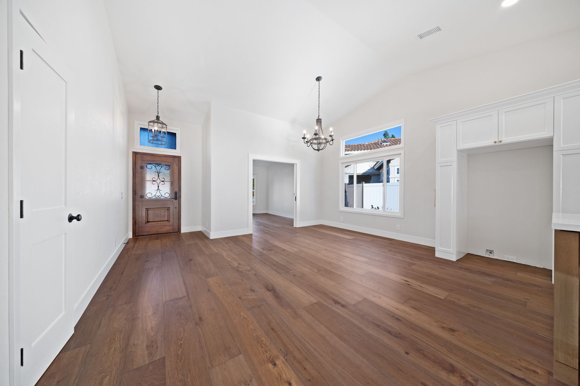 Bright empty room with hardwood floors, white walls, chandeliers, and a wooden cabinet at the far end