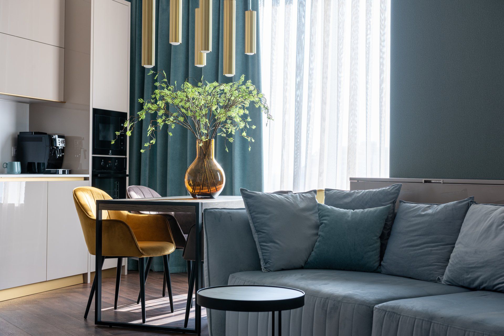 Modern living room with a gray sofa, dining table, gold chairs, and tall windows with sheer curtains.