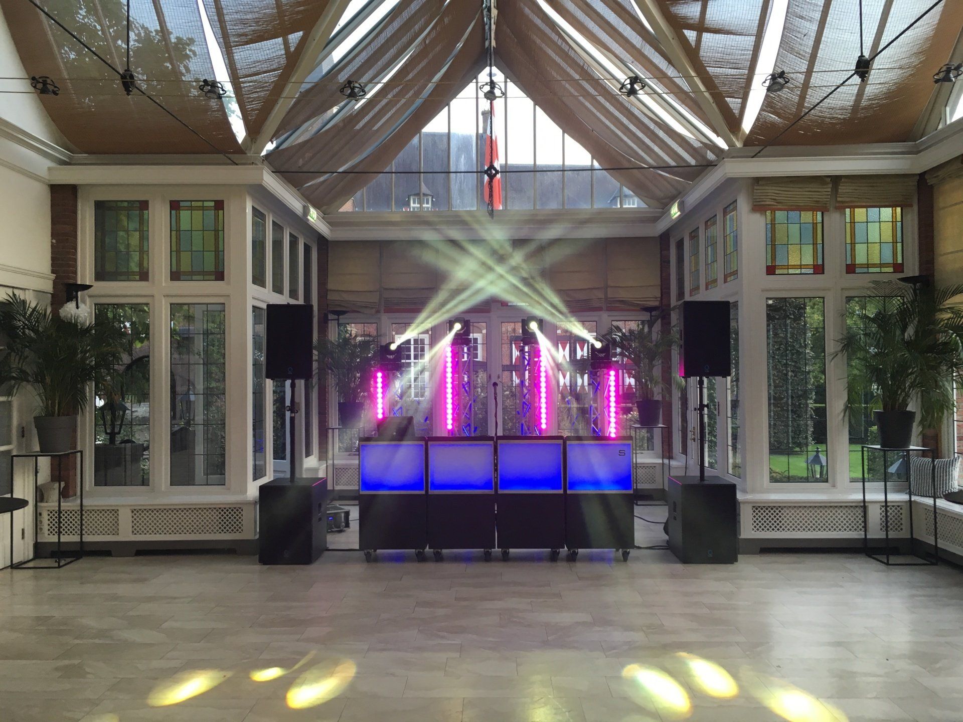 Binnenpodium met paarse en blauwe verlichting onder een hoog glazen atrium, geflankeerd door planten en luidsprekers.