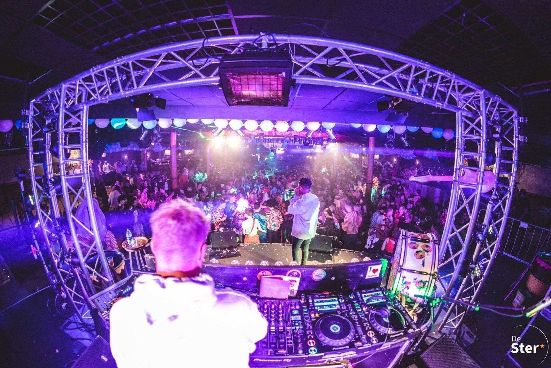 DJ op het podium voor een bomvolle zaal, met felpaarse lichten en confetti tijdens een concert.
