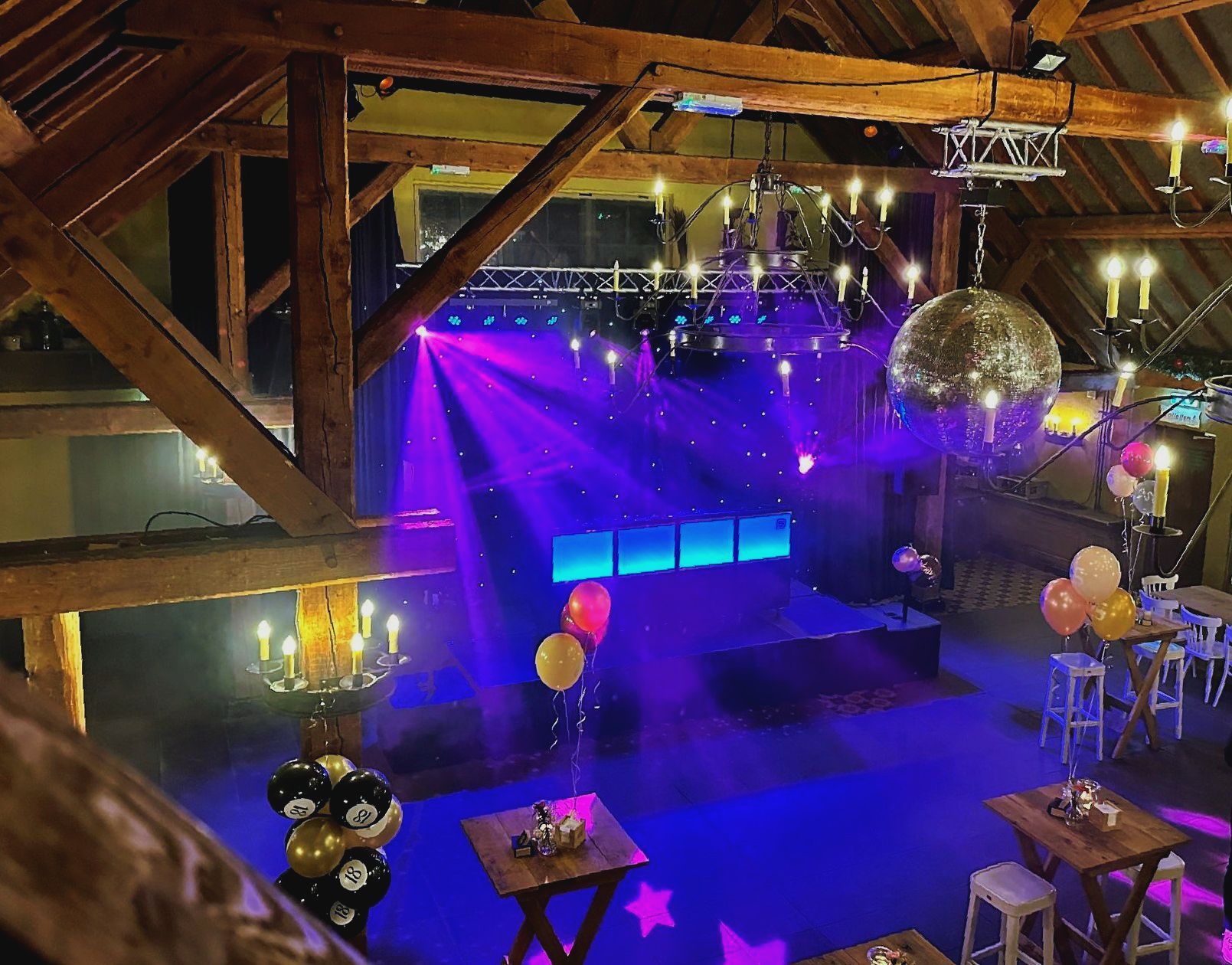 Een lege nachtclub met paarsblauwe podiumverlichting, een discobal, ballonnen en houten balken aan het plafond.