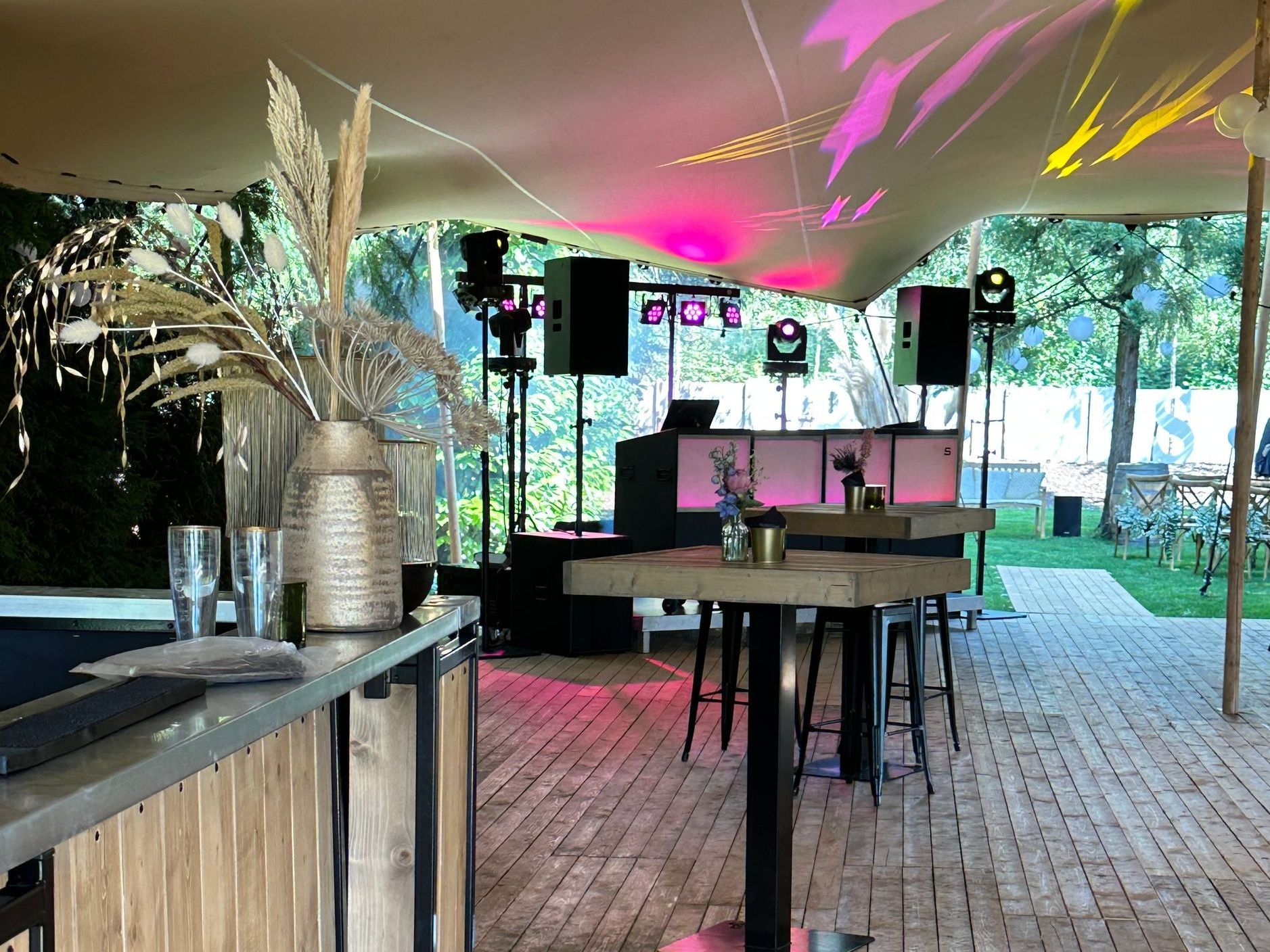 Een evenemententent in de buitenlucht met een dj-booth, speakers, roze verlichting en cocktailtafels
