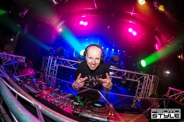 DJ in een clubbooth met kleurrijke neonlichten en een podiumachtergrond die door het publiek wordt verlicht.