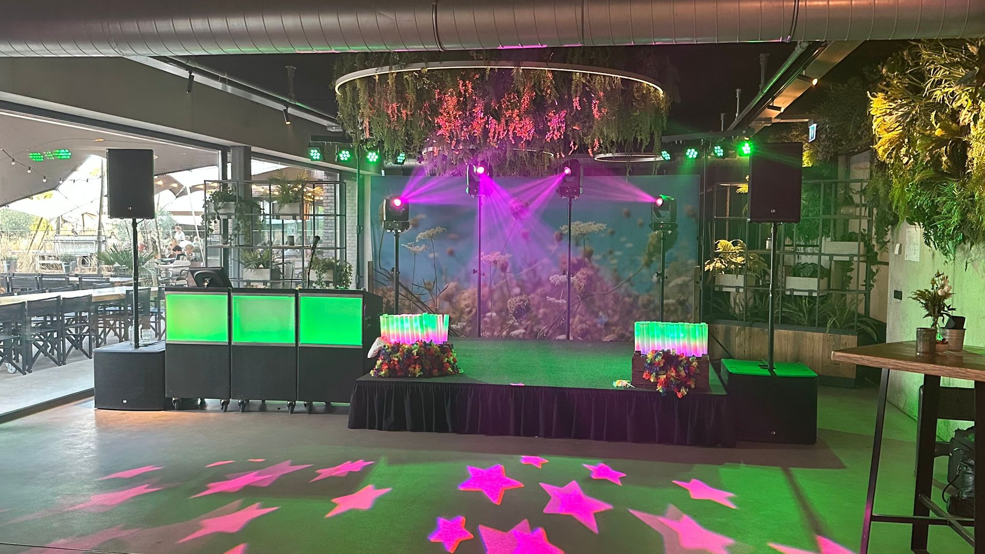 Een podium met roze en groene laserlichten, een dj-booth en lichtgevende projecties op de vloer.