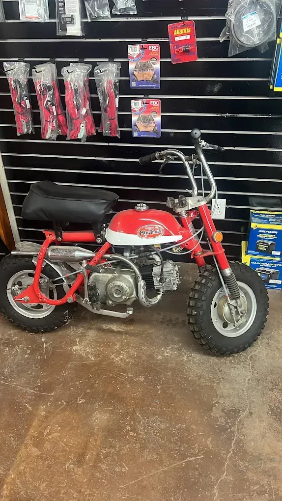Red mini motorcycle displayed in a shop, with chrome handlebars and a black seat against a wall of accessories.