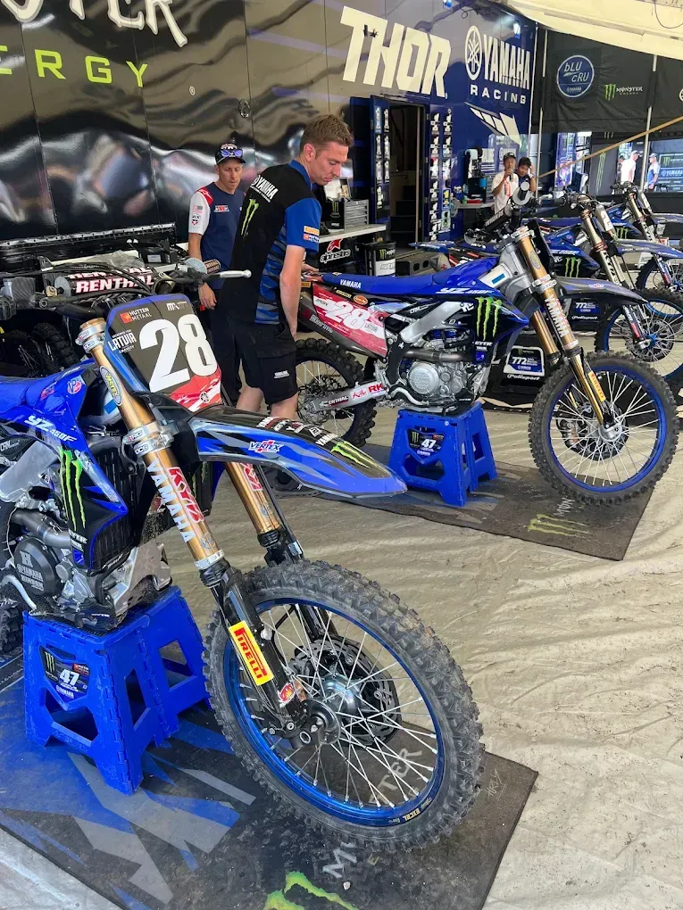 Blue dirt bikes on display in a motocross pit area, with a rider standing beside them under a Thor canopy.