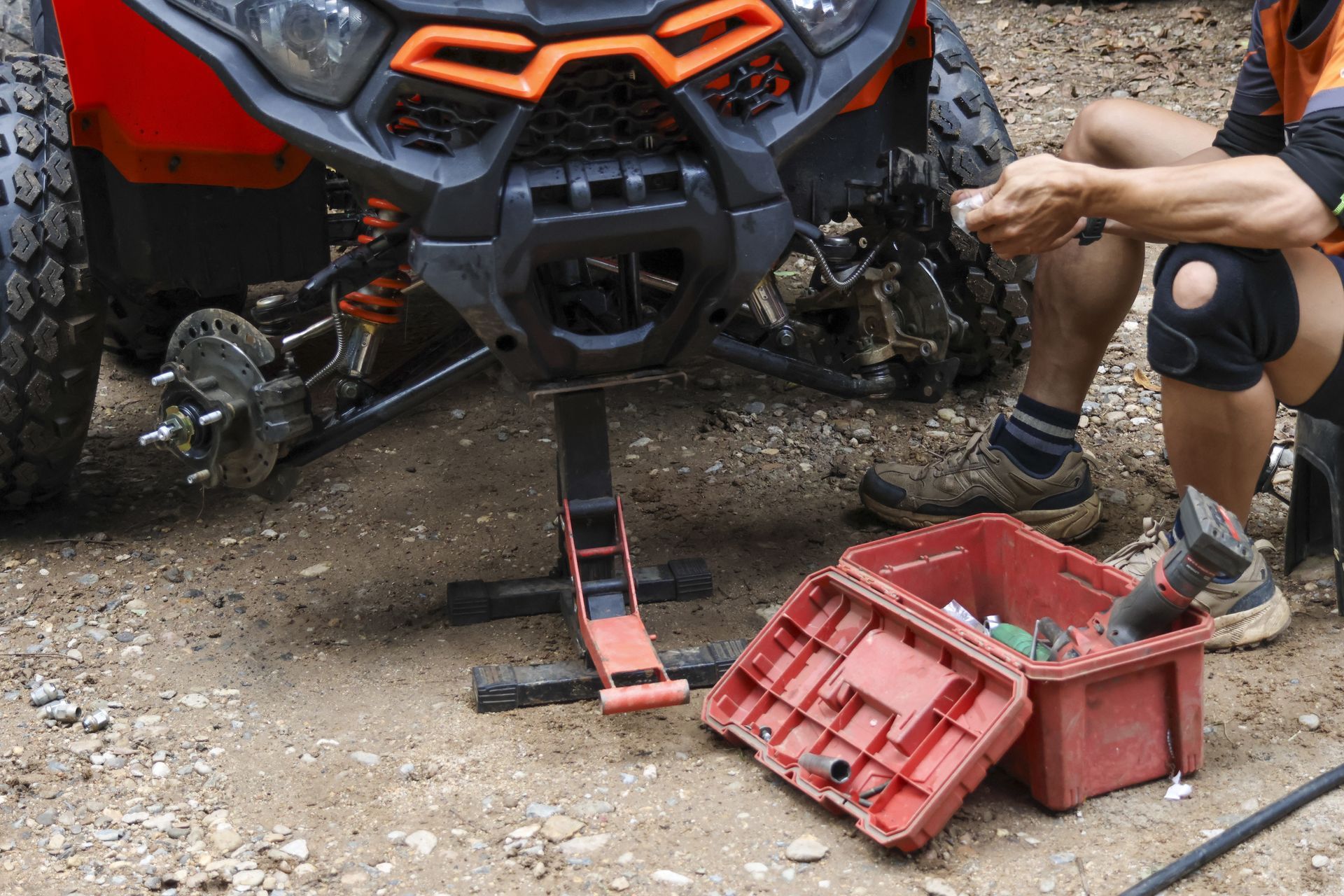 Person kneeling beside red ATV, fixing front wheel with tools on gravel trail.
