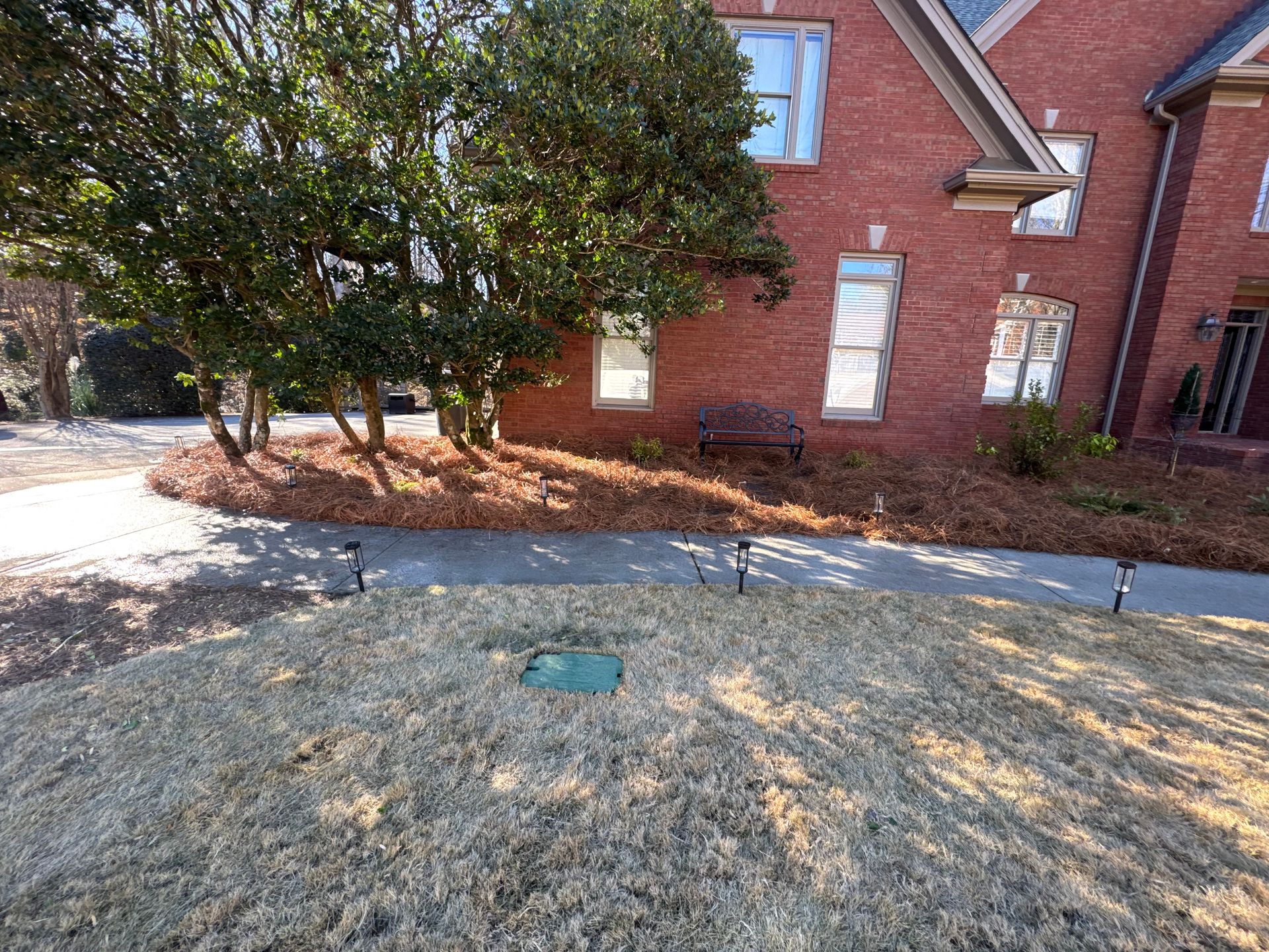 Front yard with dry grass and pathway, flanked by brick house and leafy trees. A metal bench sits near the building.
