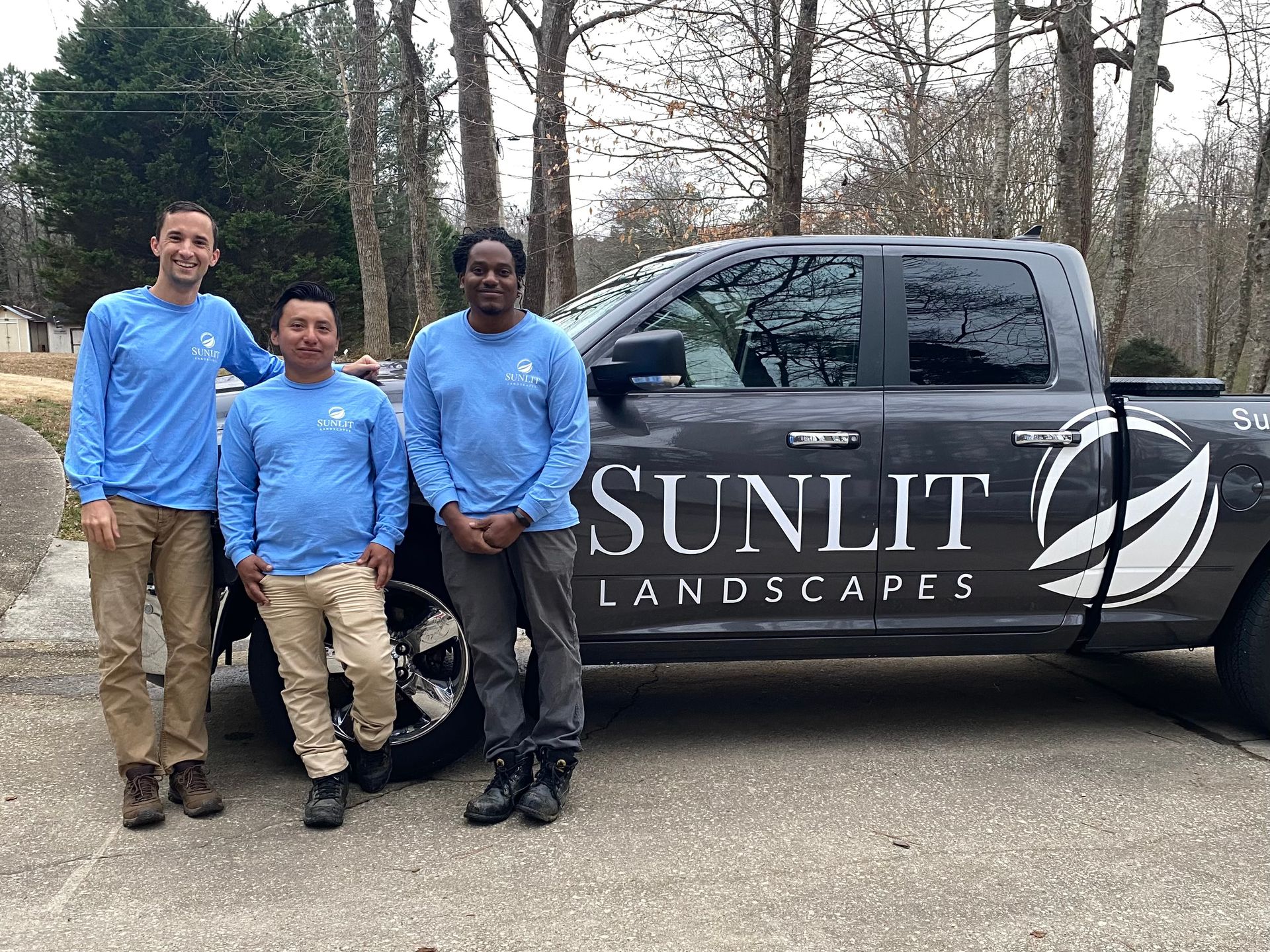 Three people in light blue long-sleeve shirts stand smiling next to a gray Sunlit Landscapes pickup truck outdoors.