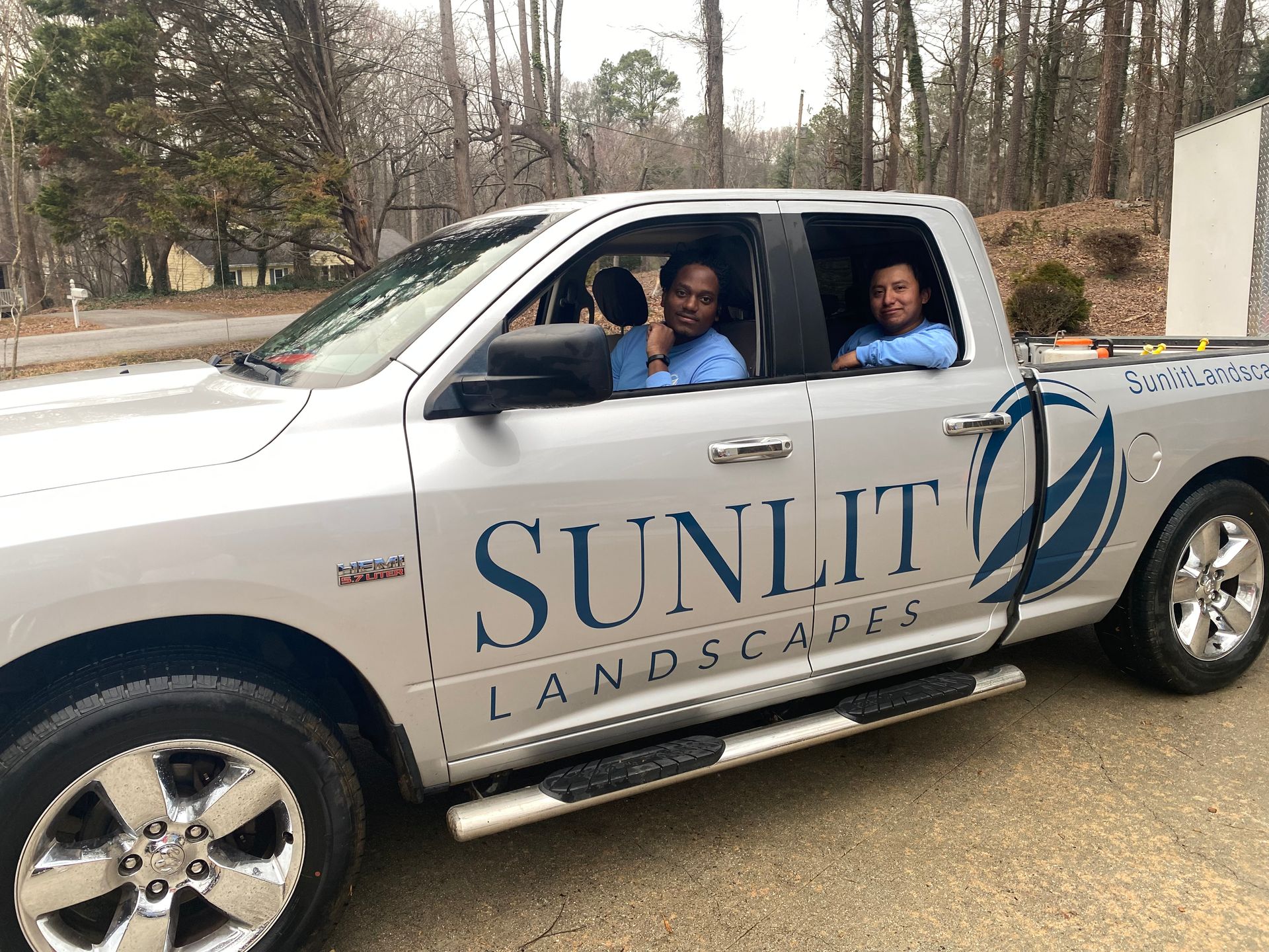 Two people in light blue shirts sit in a silver Sunlit Landscapes pickup truck parked outdoors.
