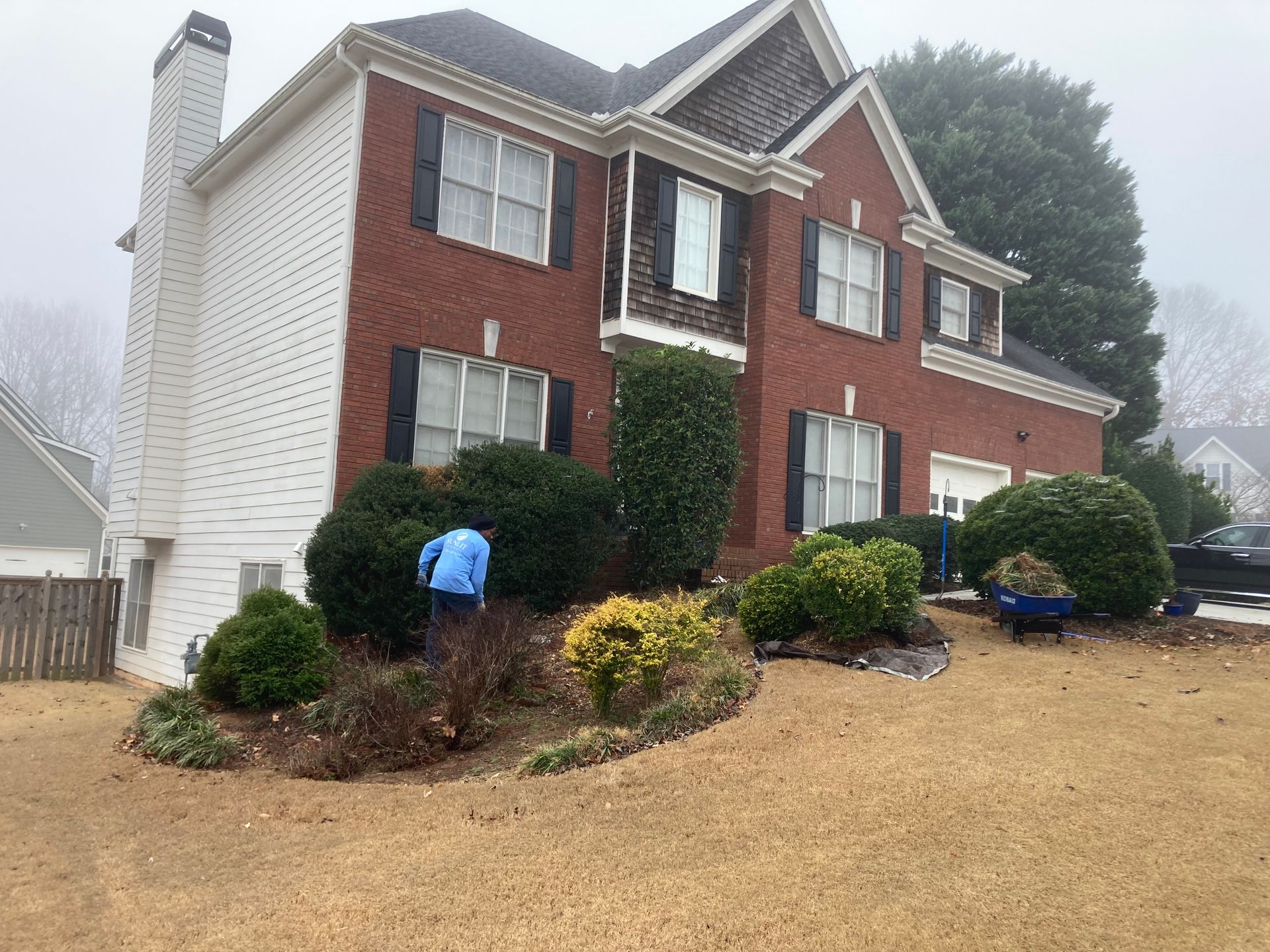 A person in blue works on landscaping near a large, two-story red brick house with white trim.